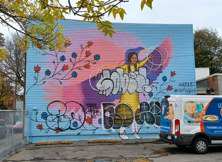La murale occupe un pan de mur entier et illustre une figure féminine devant une silouhette d'aigle. Des cerceaux colorés reposent sur ses bras et l'arrière-plan de la murale est peint en bleu ciel, violet et rose. Des graffitis sont dispersés sur la murale.