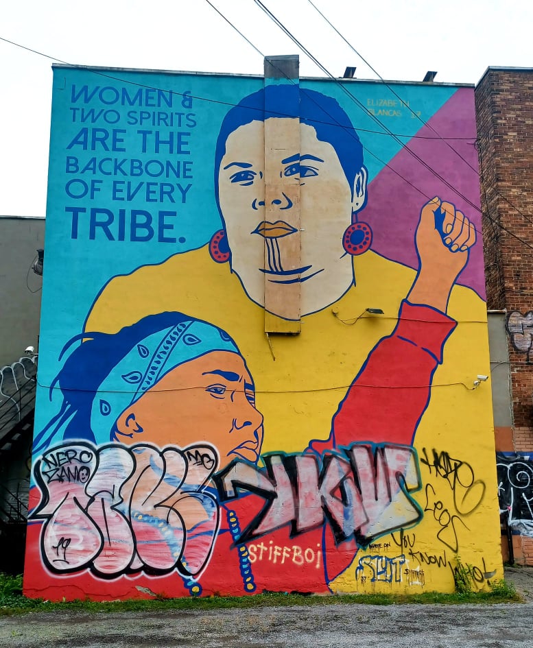 La murale de Blancas en 2021, des graffitis recouvrent le bas de l'œuvre. Sur la façade d'un immeuble, deux figures monumentales sont représentées sur un fond cyan et violet, où la phrase Women & Two Spirits are the Backbone of Every Tribe est inscrite dans le coin supérieur gauche.