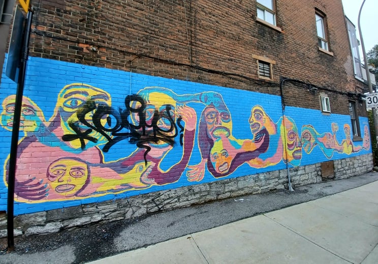 œuvre par Cedar Eve Peters, prise en photo en 2021, où un graffiti couvre une partie à droite de la murale. Sur un fond bleu ciel, diverses figures colorées mi-humaines, mi-animales sont représentées le long d'un bâtiment.